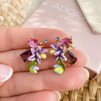 Detalle de pendientes florales esmaltados con cristal morado sostenidos en una mano con manicura natural.